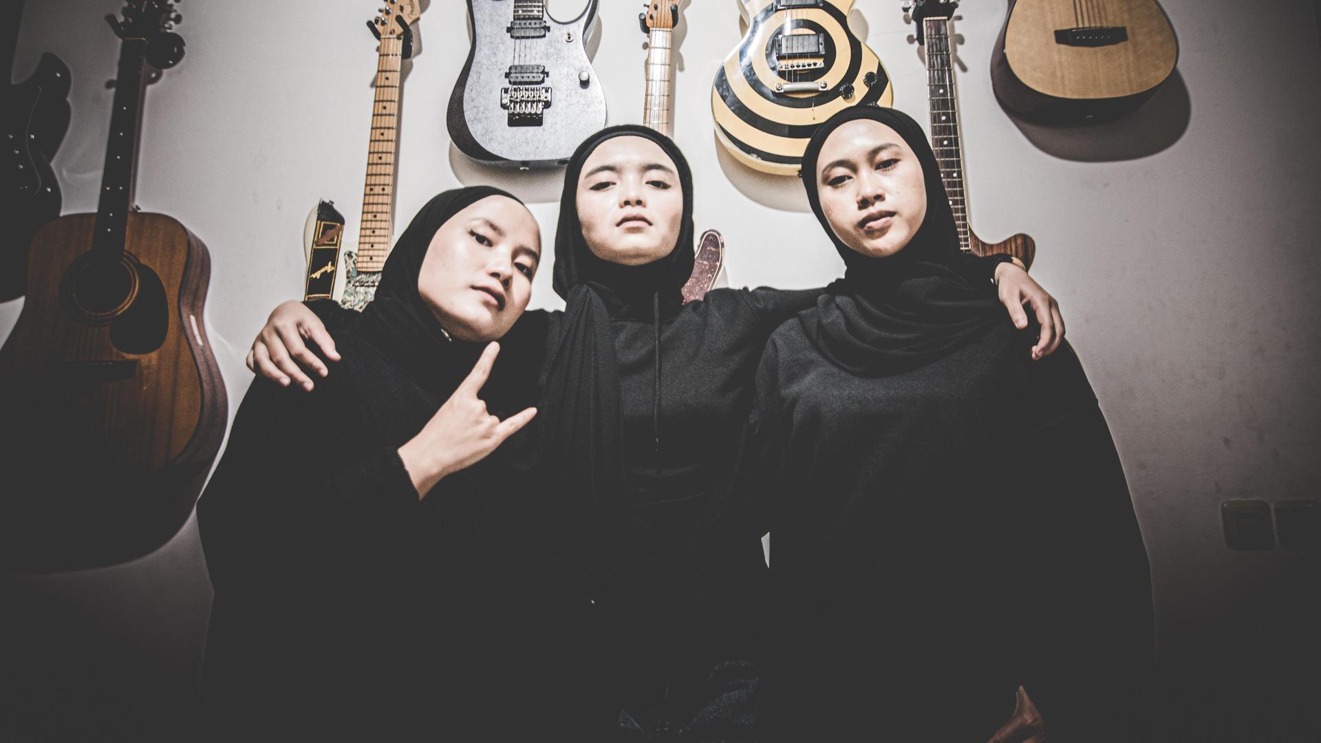 Die indonesische Metalband "Voice of Baceprot". Drei junge indonesische Frauen mit Kopftüchern stehen vor Gitarren, die an der Wand hängen. Die Frau links macht die "Pommesgabel", die in der Mitte hat die Arme um die beiden anderen gelegt.