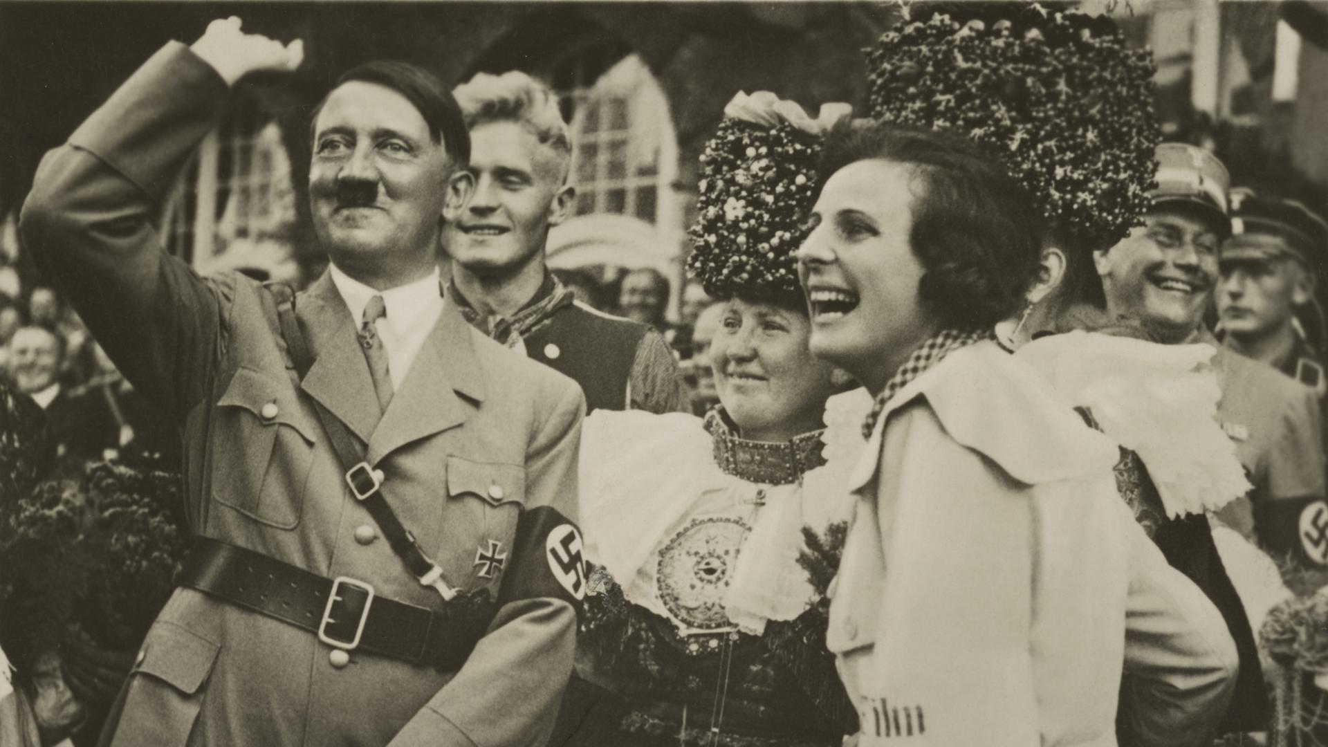 Eine Schwarzweiß-Fotografie zeigt Adolf Hitler mit erhoben und angewinkeltem rechtem Arm neben einer ihn anlachenden Frau vor einer Reihe in Trachten gekleideter Frauen.
