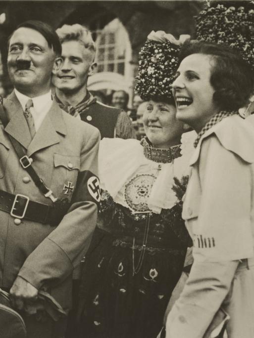 Eine Schwarzweiß-Fotografie zeigt Adolf Hitler mit erhoben und angewinkeltem rechtem Arm neben einer ihn anlachenden Frau vor einer Reihe in Trachten gekleideter Frauen.