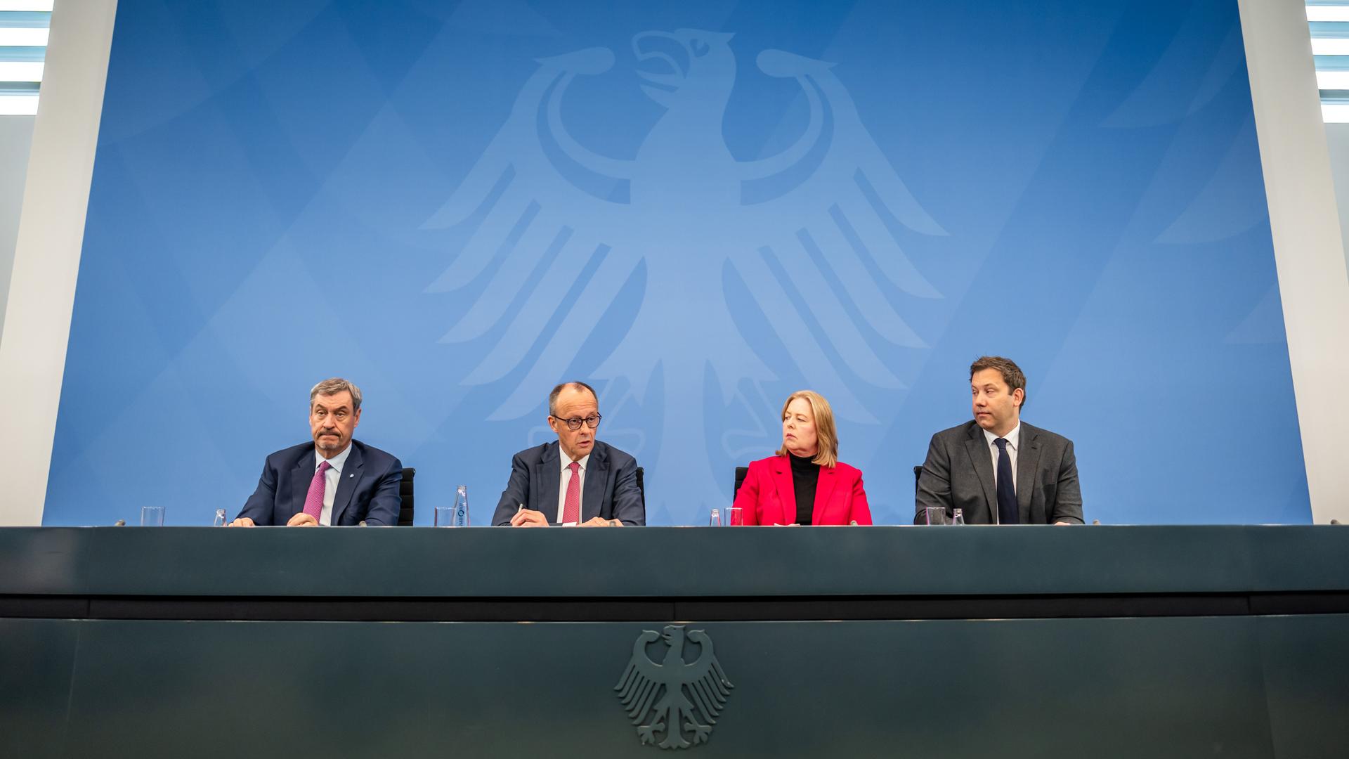 Markus Söder (CSU), Friedrich Merz (CDU), Bärbel Bas (SPD) sowie Lars Klingbeil (SPD) nehmen an einer Pressekonferenz teil und sitzen an einem schwarzen Pult vor blauer Wand.