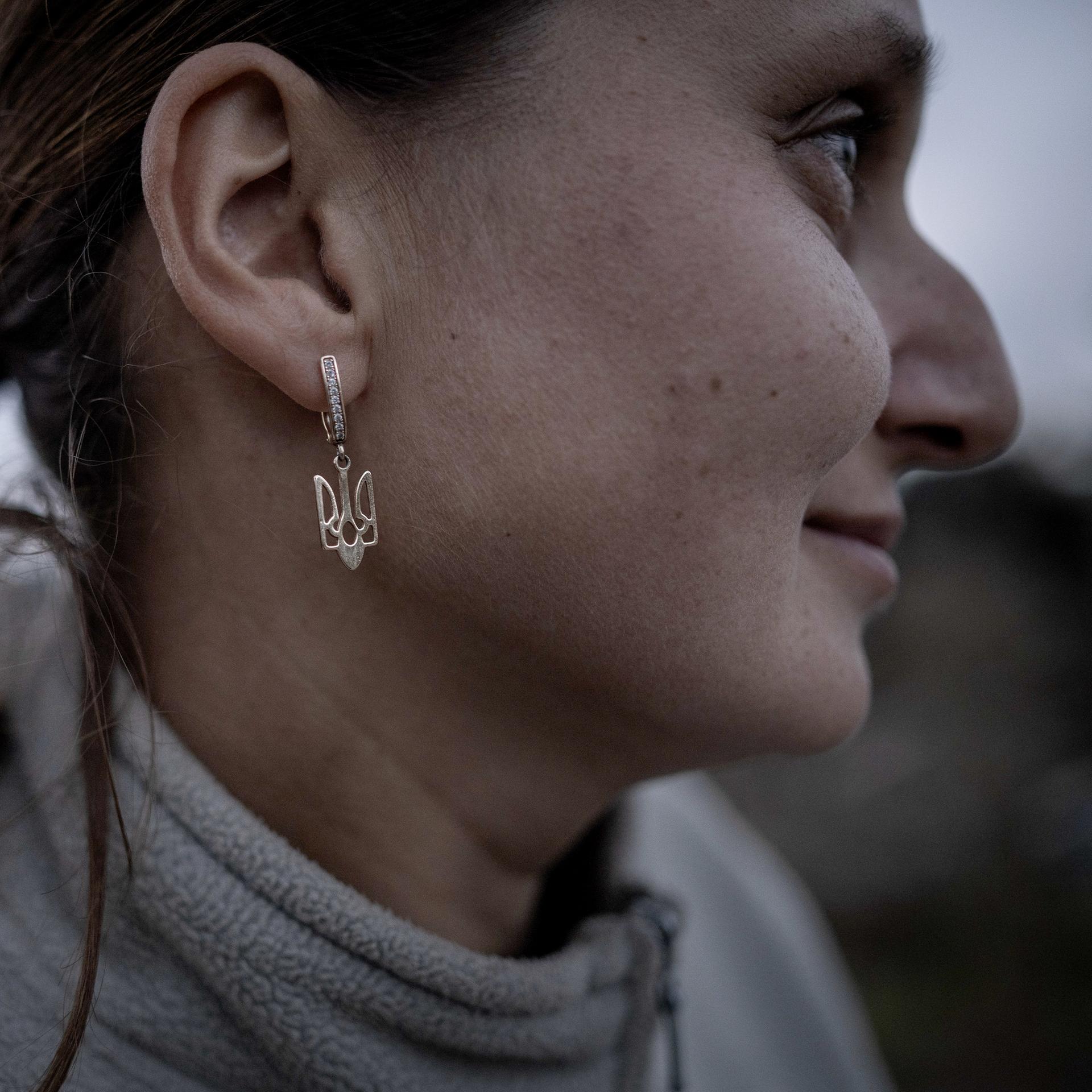 Nahaufnahme einer Frau im Profil, die als Ohrringe silberne Anhänger in Form des ukrainischen Wappens trägt.