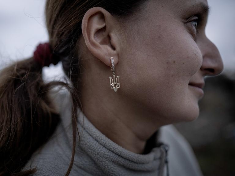 Nahaufnahme einer Frau im Profil, die als Ohrringe silberne Anhänger in Form des ukrainischen Wappens trägt.