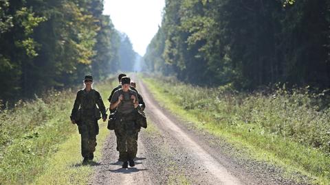 Drei uniformierte Frauen mit militärischer Ausrüstung auf einer Straße, die durch den Wald führt