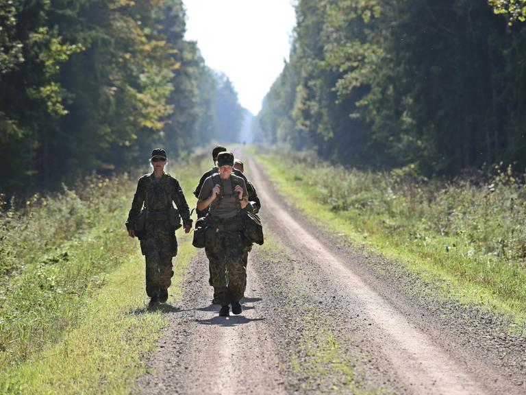 Drei uniformierte Frauen mit militärischer Ausrüstung auf einer Straße, die durch den Wald führt