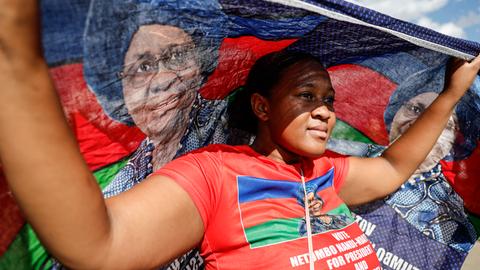 Eine junge Frau trägt während einer Wahlkampfveranstaltung eine Flagge mit dem Gesicht von Netumbo Nandi-Ndaitwah. Ihre Kleidung hat die Farben der namibischen Flagge: blau, rot, grün.