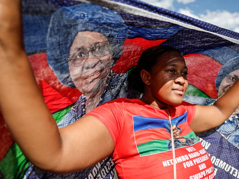 Eine junge Frau trägt während einer Wahlkampfveranstaltung eine Flagge mit dem Gesicht von Netumbo Nandi-Ndaitwah. Ihre Kleidung hat die Farben der namibischen Flagge: blau, rot, grün.