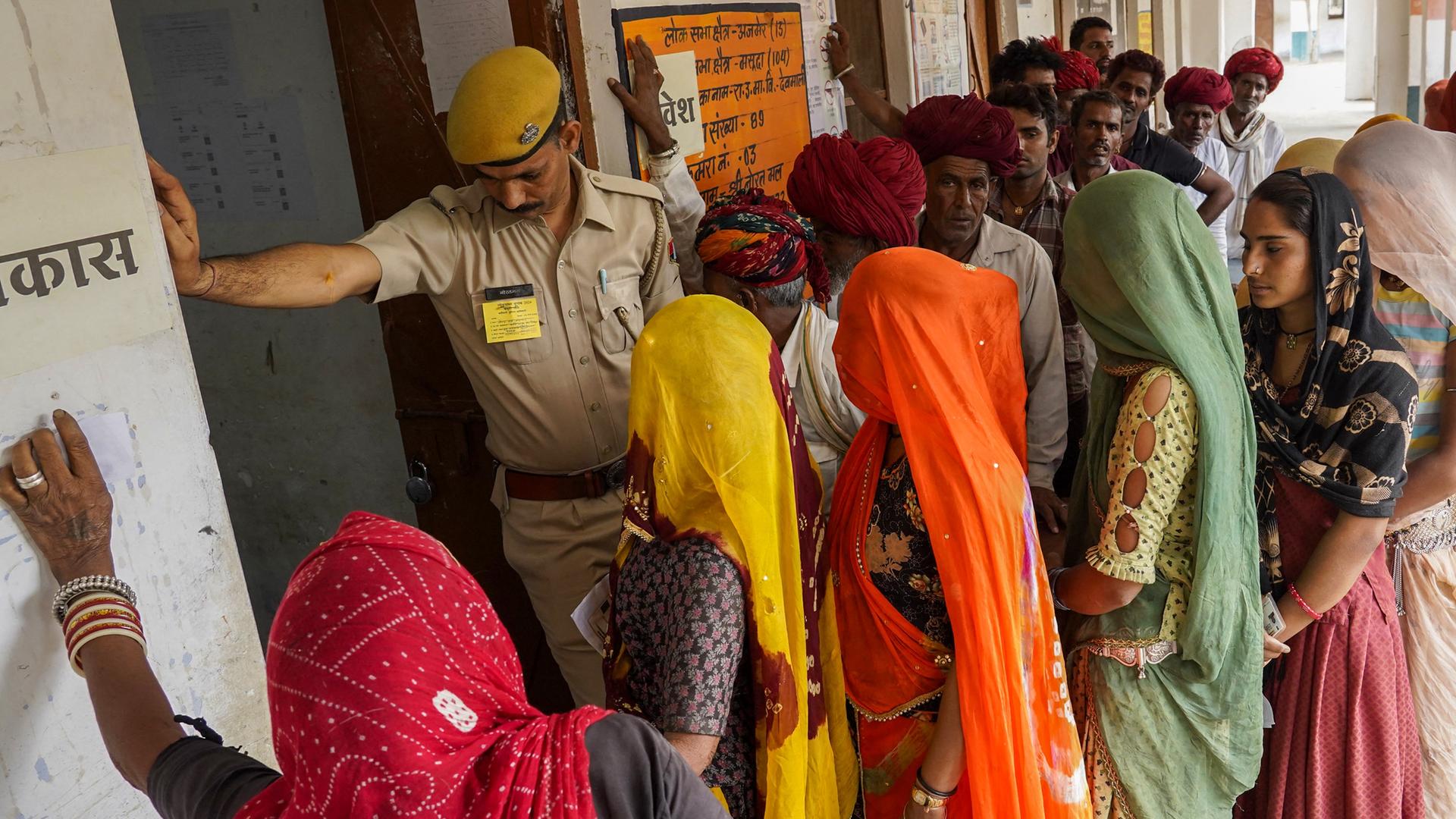 Wähler stehen vor einer Stimmabgabestation in Ajmer in Indien. Ein Beamter hält einen Arm vor die Tür, damit die Leute einzeln eintreten.