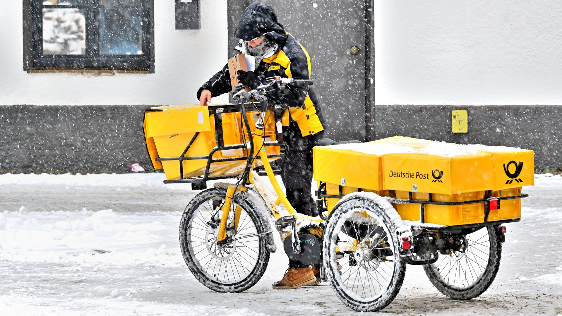 Eine Postzustellerin mit ihrem gelben Lastenfahrrad an einem schneereichen Wintertag in Bad Reichenhall bei der Arbeit. Sie trägt im Schneetreiben die Post aus.