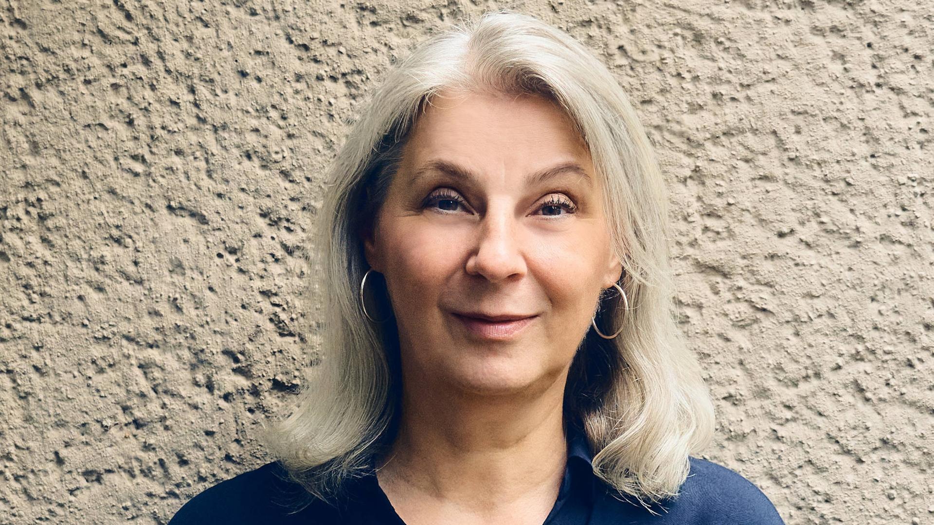 Ruth Olshan lächelt in die Kamera. Sie trägt eine blaue Bluse und kinnlanges graues Haar. Ruth Olshan lächelt in die Kamera. Sie trägt eine blaue Bluse und kinnlanges graues Haar.