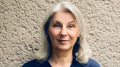 Ruth Olshan lächelt in die Kamera. Sie trägt eine blaue Bluse und kinnlanges graues Haar. Ruth Olshan lächelt in die Kamera. Sie trägt eine blaue Bluse und kinnlanges graues Haar.