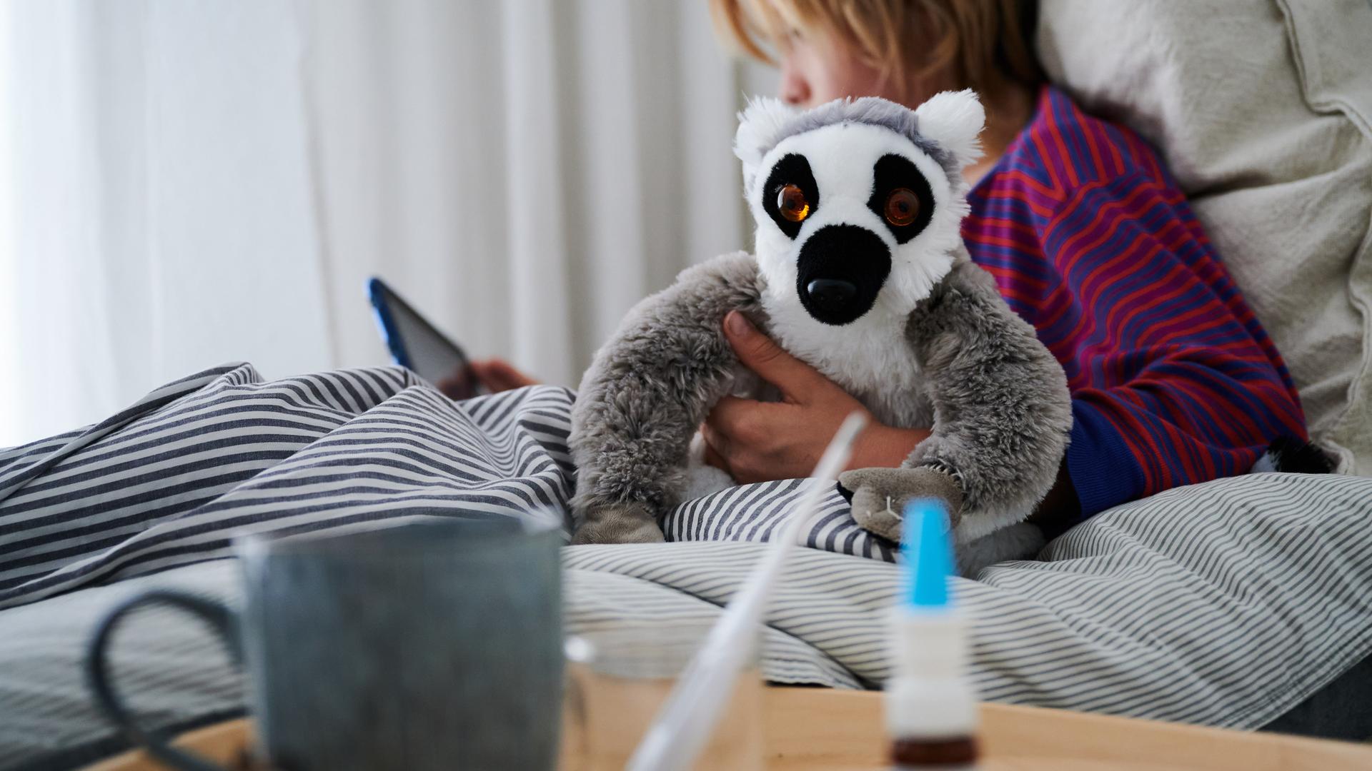 Fieberthermometer, Nasenspray und eine Tasse Tee stehen vor einem Bett, in dem ein Kind mit Kuscheltier auf ein Tablet guckt.