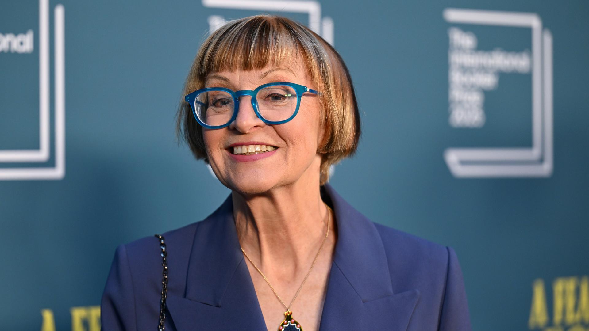 Philippa Perry steht vor einer blauen Fotowand und schaut lächelnd in die Kamera. Sie trägt eine Brille mit blauen Rahmen. Philippa Perry steht vor einer blauen Fotowand und schaut lächelnd in die Kamera. Sie trägt eine Brille mit blauen Rahmen.