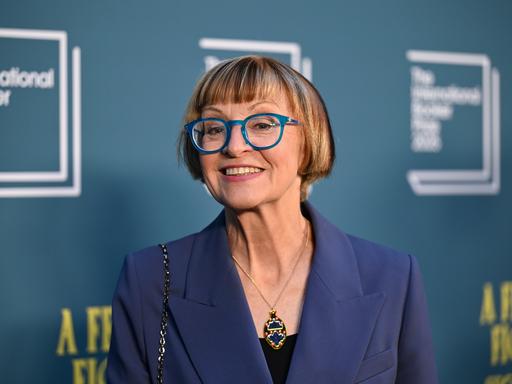 Philippa Perry steht vor einer blauen Fotowand und schaut lächelnd in die Kamera. Sie trägt eine Brille mit blauen Rahmen.