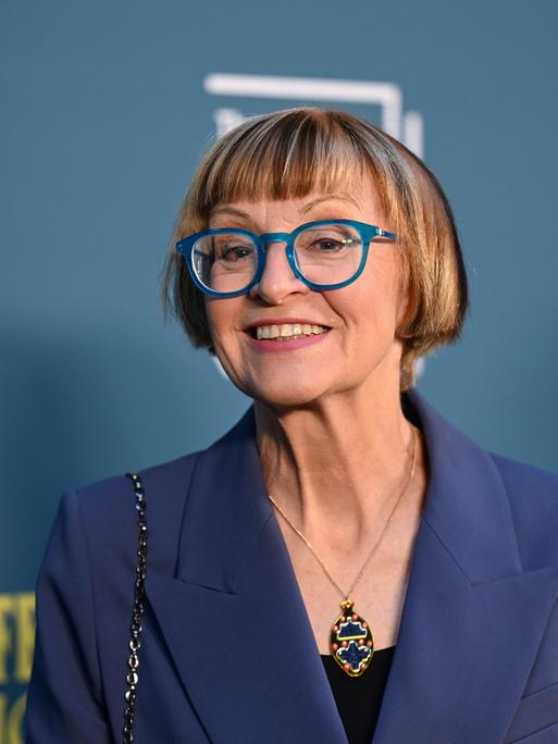 Philippa Perry steht vor einer blauen Fotowand und schaut lächelnd in die Kamera. Sie trägt eine Brille mit blauen Rahmen. Philippa Perry steht vor einer blauen Fotowand und schaut lächelnd in die Kamera. Sie trägt eine Brille mit blauen Rahmen.
