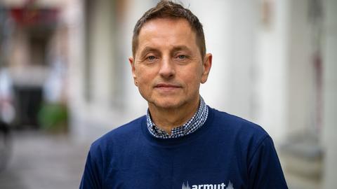 Thomas de Vachroi lächelt in die Kamera. Er trägt ein blaues Shirt mit der Aufschrift "Armut eine Stimme geben". Thomas de Vachroi lächelt in die Kamera. Er trägt ein blaues Shirt mit der Aufschrift "Armut eine Stimme geben".