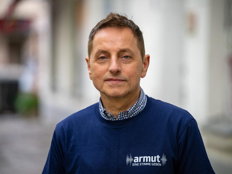 Thomas de Vachroi lächelt in die Kamera. Er trägt ein blaues Shirt mit der Aufschrift "Armut eine Stimme geben".