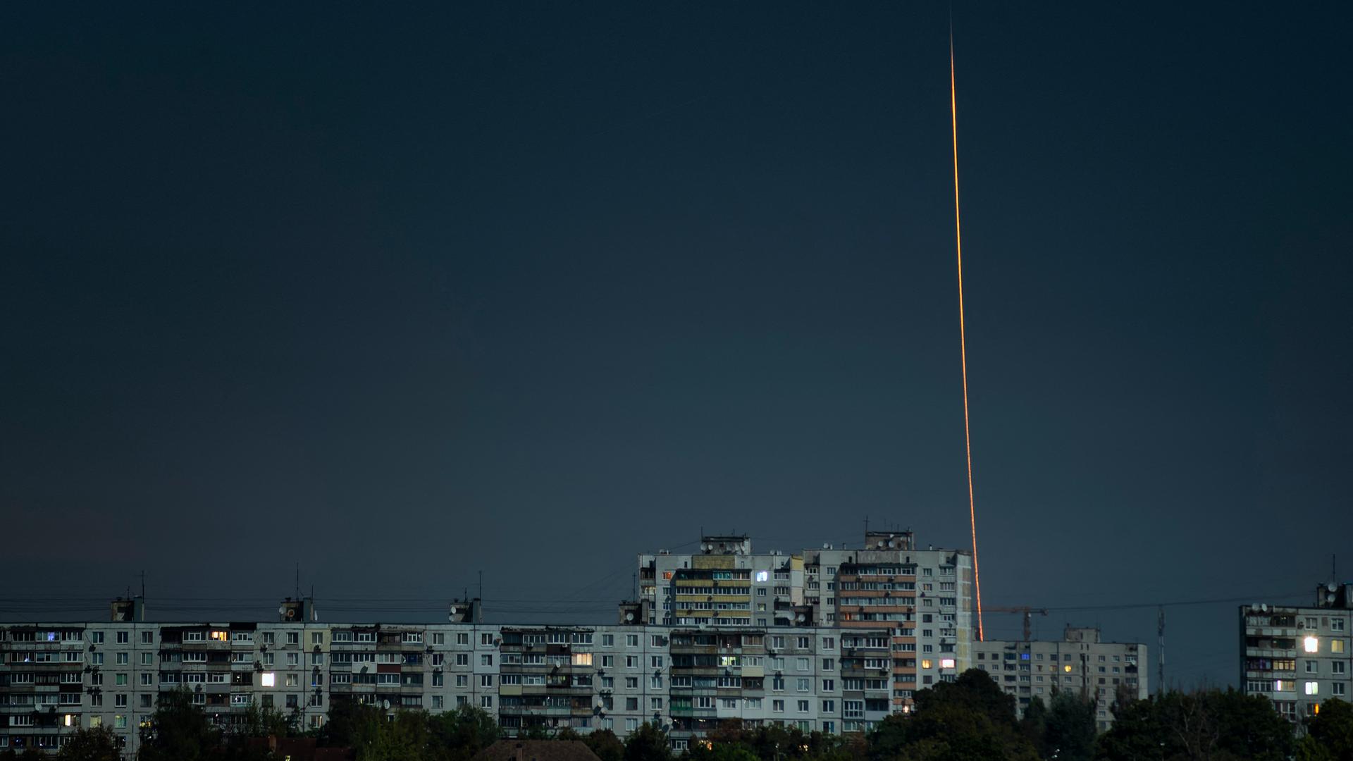 Über einer dunklen Stadt zieht eine gestartete Rakete einen hellen Streifen hinter sich her.