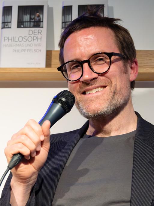 Ein braunhaariger Mann mit runder Brille und kurzem Bart sitzt vor einem Bücherregal und hält ein Mikrofon in der Hand. Er lächelt in die Kamera. Er trägt ein Nadelstreifenjackett und ein graues T-Shirt. 