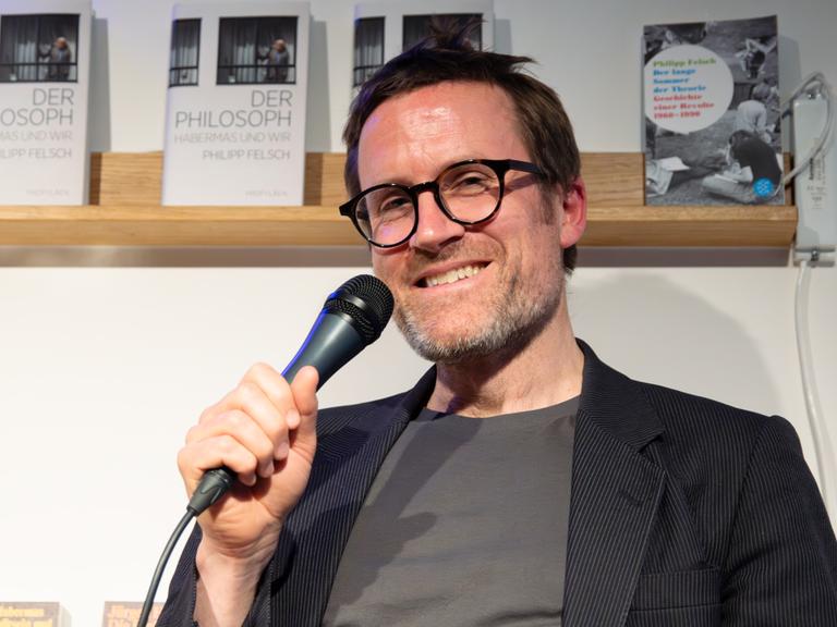 Ein braunhaariger Mann mit runder Brille und kurzem Bart sitzt vor einem Bücherregal und hält ein Mikrofon in der Hand. Er lächelt in die Kamera. Er trägt ein Nadelstreifenjackett und ein graues T-Shirt. 