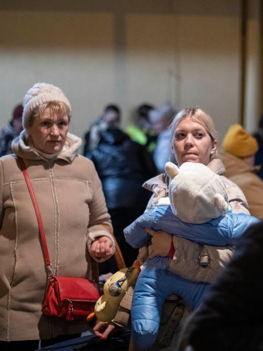 Zwei weiße, blonde Frauen in Winterkleidung stehen zwischen Gepäck und anderen Frauen an vermutlich einem Bahnhof. Die rechte der beiden Frauen hat ein kleines Kind auf dem Arm.