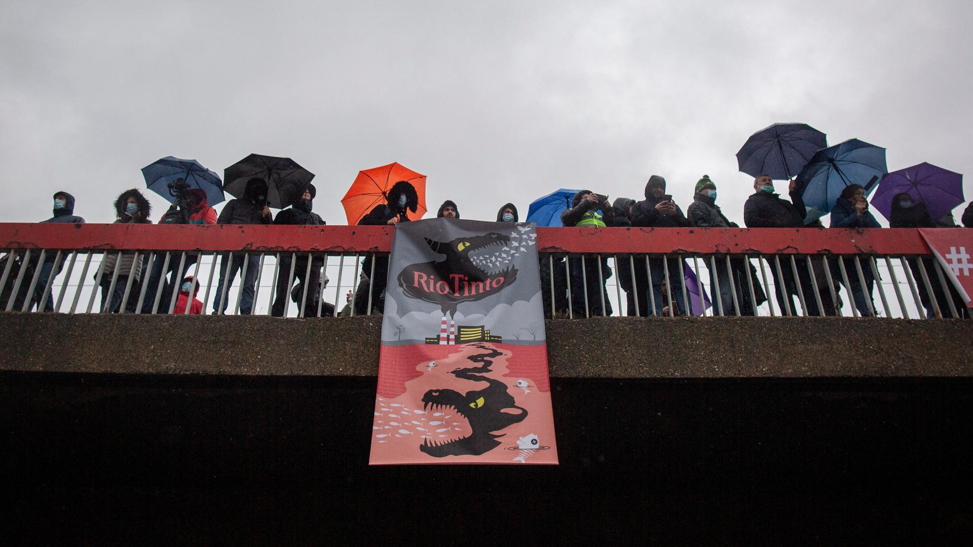 Demonstranten auf einer Brücke halten ein Banner, auf dem sich ein illustriertes Monster ausgehend von einem Fabrikgebäude in die Erde frisst.