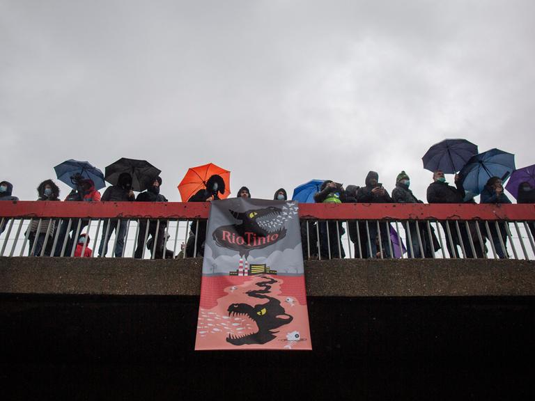 Demonstranten auf einer Brücke halten ein Banner, auf dem sich ein illustriertes Monster ausgehend von einem Fabrikgebäude in die Erde frisst.