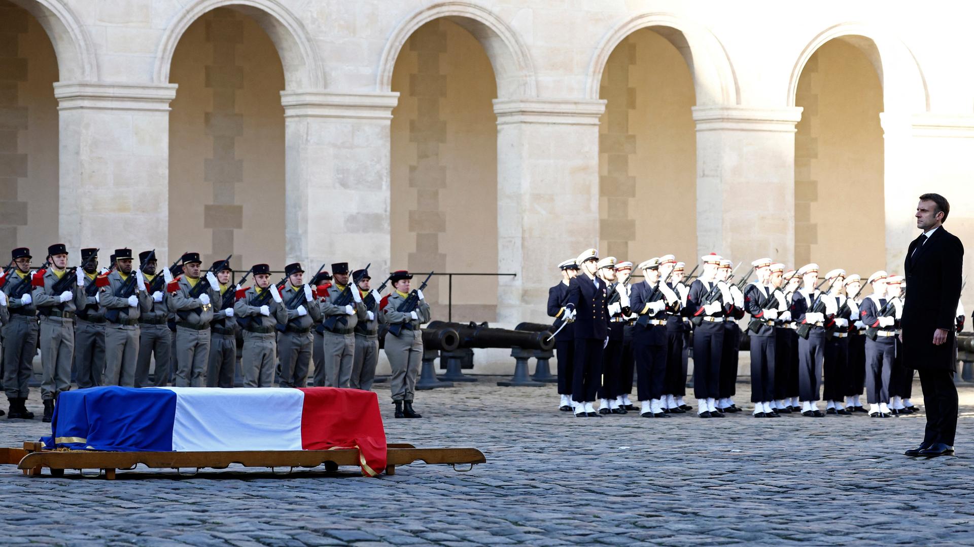 Macron steht vor dem mit einer Frankreich-Flagge bedeckten Sarg, der auf dem Boden steht. Dahinter Soldaten und ein Musikcorps in Reih und Glied.