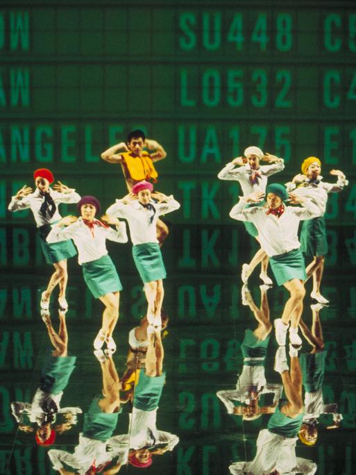 Sieben Personen, die an Flugbegleiter erinnen, stehen vor einer Leinwand. An der Leinwand sind in großen Buchstaben Flugnummern und Ziele angeschlagen. Das Bild ist Teil einer Videoinstallation eines japanischen Künstlerkollektivs.