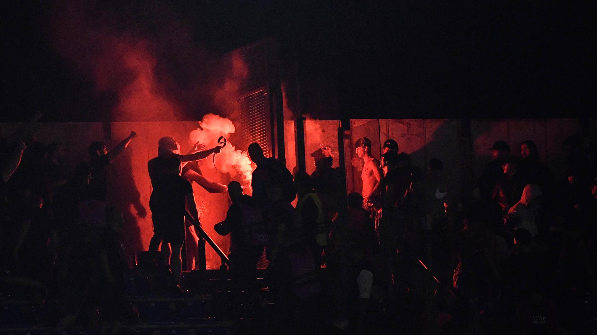 Auf einer Tribüne stehen dunkel gekleidete Menschen. Pyrotechnik brennt rot.