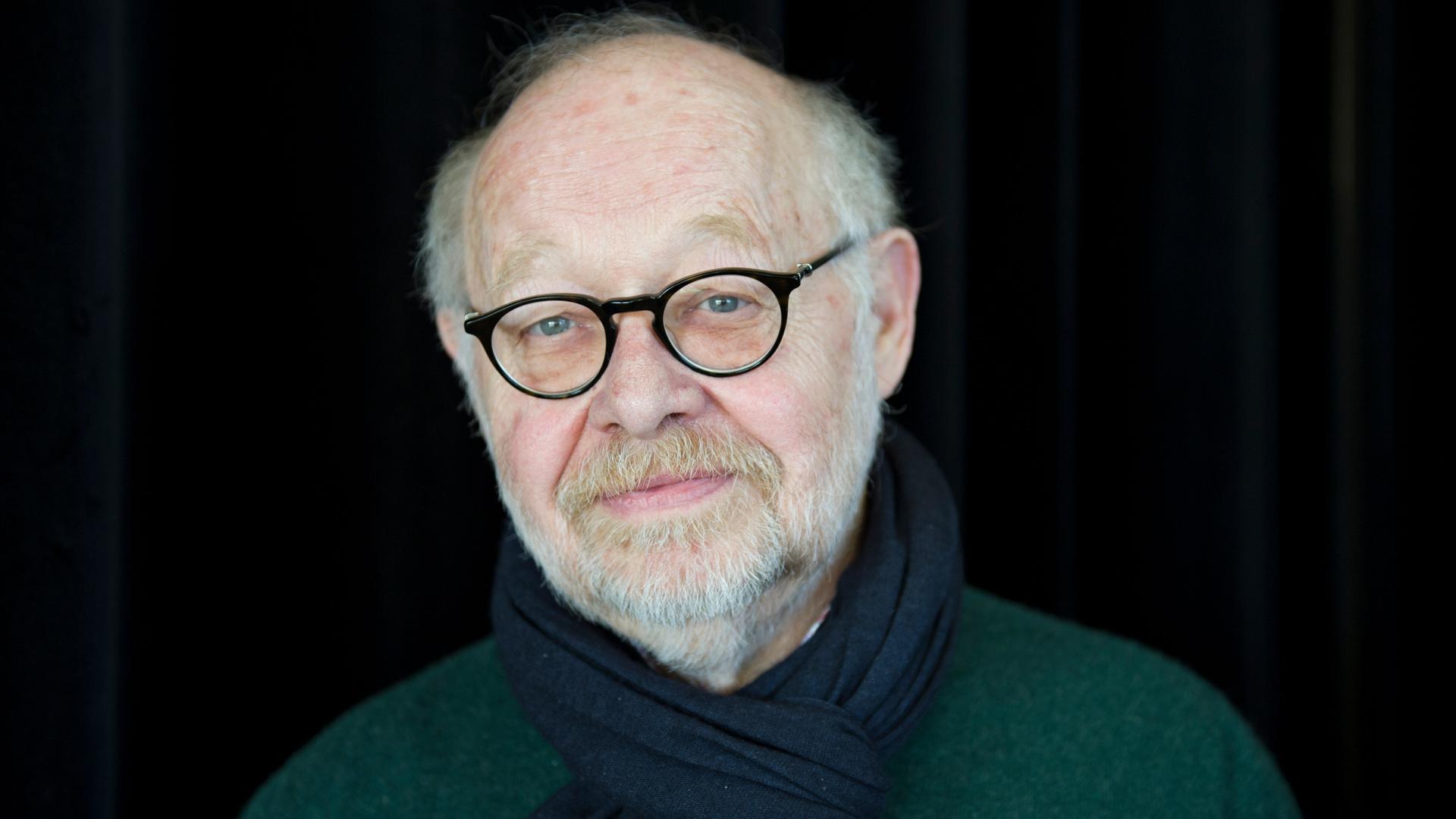 Der Regisseur Jürgen Flimm, mit Brille und weißem Bart, steht vor einem dunklen Hintergrund und schaut freundlich konzentriert in die Kamera.
