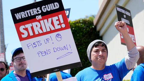Mehrere Demonstranten halten in Hollywood Schilder mit der Aufschrift "Streik" in die Höhe, im Vordergrund reckt einer von ihnen die Faust in die Höhe. 