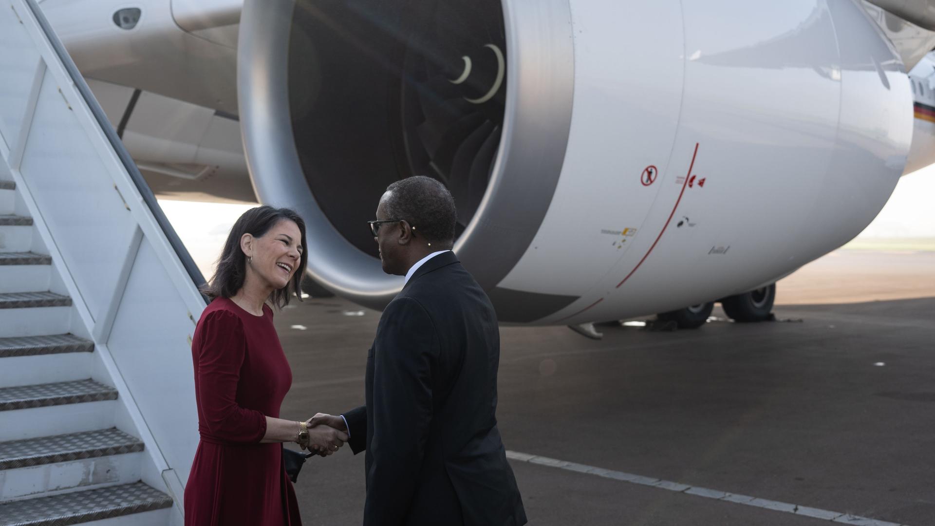 Außenministerin Annalena Baerbock wird in Ruanda von ihrem Amtskollegen Vincent Biruta begrüßt. Sie stehen vor einer Passagiermaschine, im Hintergrund sind die Gangway und ein Triebwerk zu sehen.
