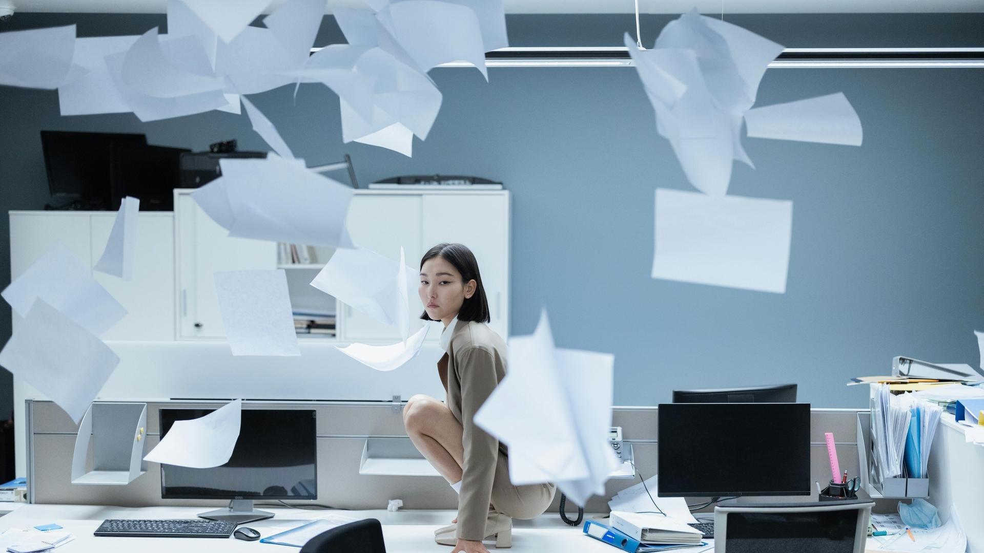 Eine Frau in einem Büro, um sie herum fliegt Papier herum.