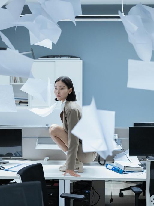 Eine Frau in einem Büro, um sie herum fliegt Papier herum.