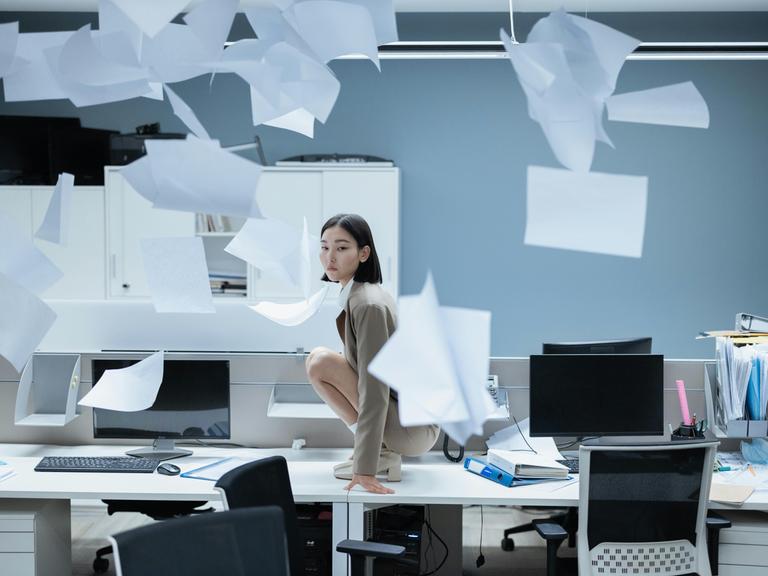 Eine Frau in einem Büro, um sie herum fliegt Papier herum.