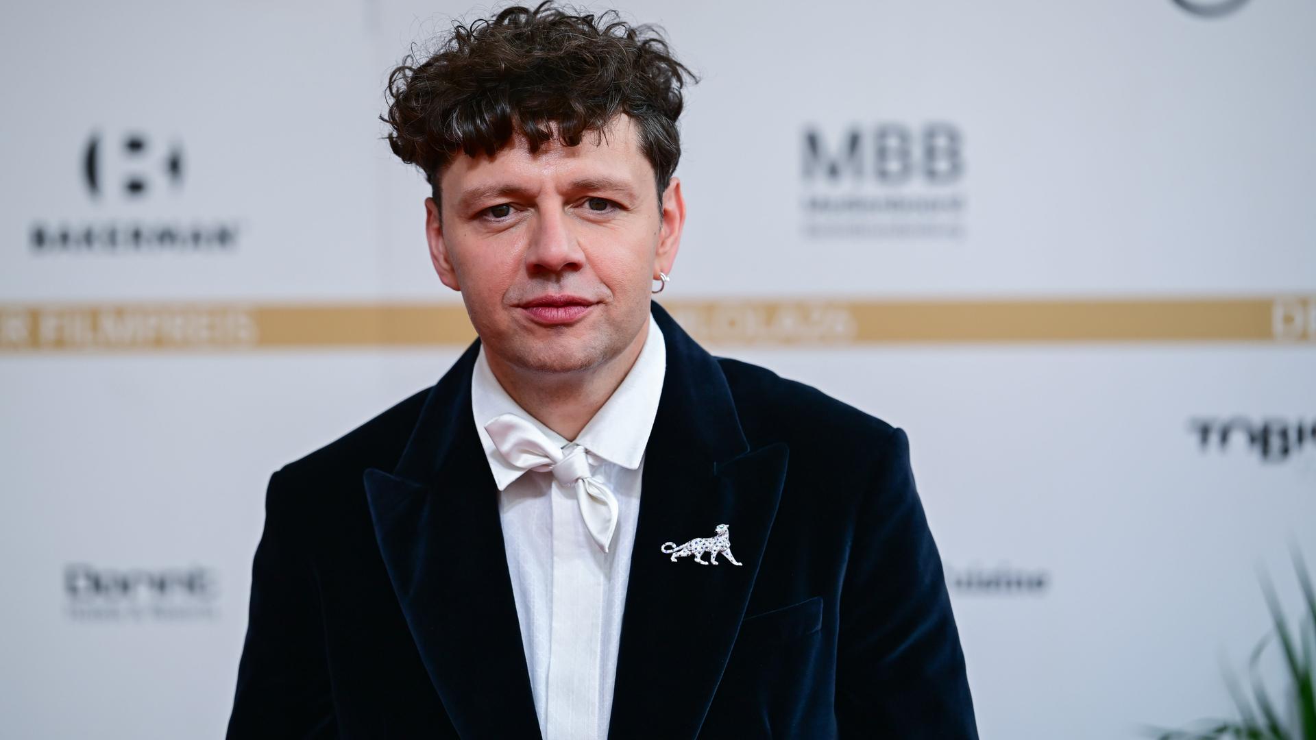 Ein Mann, der Schauspieler und Musiker Christian Friedel, seht in schwarzem Anzug mit weißer Fliege vor einem weißen Hintergrund mit unscharfen Logos Ein Mann, der Schauspieler und Musiker Christian Friedel, seht in schwarzem Anzug mit weißer Fliege vor einem weißen Hintergrund mit unscharfen Logos