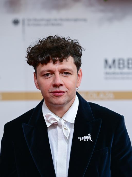 Ein Mann, der Schauspieler und Musiker Christian Friedel, seht in schwarzem Anzug mit weißer Fliege vor einem weißen Hintergrund mit unscharfen Logos Ein Mann, der Schauspieler und Musiker Christian Friedel, seht in schwarzem Anzug mit weißer Fliege vor einem weißen Hintergrund mit unscharfen Logos