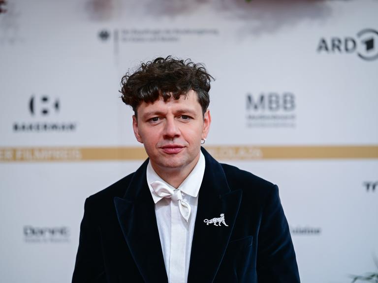 Ein Mann, der Schauspieler und Musiker Christian Friedel, seht in schwarzem Anzug mit weißer Fliege vor einem weißen Hintergrund mit unscharfen Logos Ein Mann, der Schauspieler und Musiker Christian Friedel, seht in schwarzem Anzug mit weißer Fliege vor einem weißen Hintergrund mit unscharfen Logos