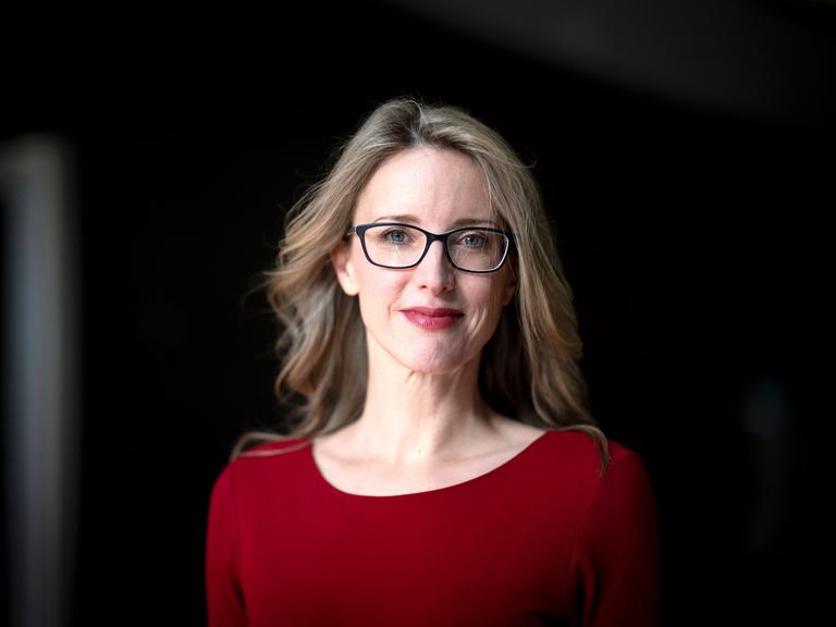 Alena Buyx posiert mit rotem Kleid und schwarzer Brille vor einem dunklen Hintergrund für ein Pressefoto.