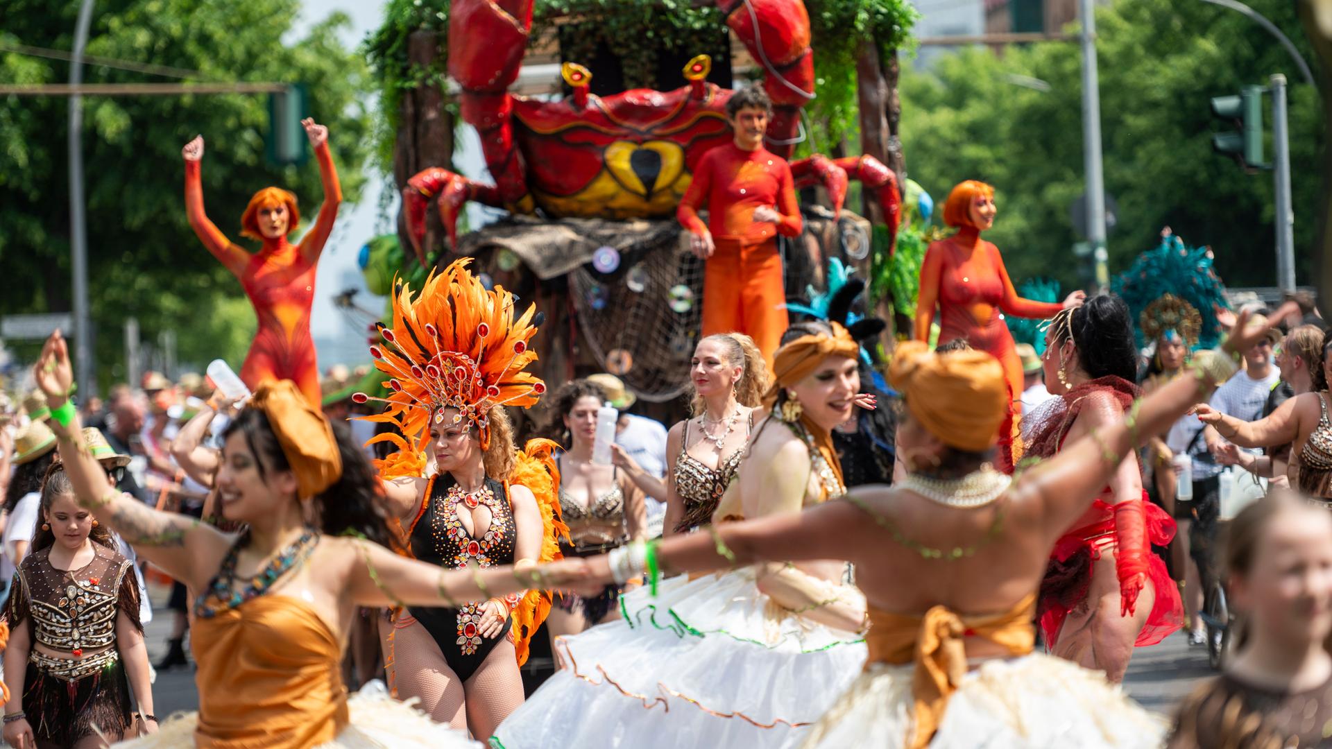 Auf einer Straßenparade tanzen Menschen in bunten Kostümen, im Hintergrund ist ein Umzugswagen zu sehen.
