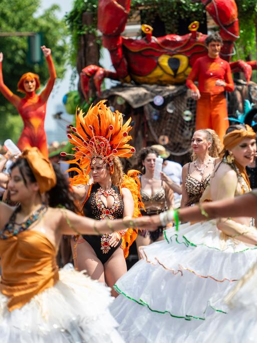 Auf einer Straßenparade tanzen Menschen in bunten Kostümen, im Hintergrund ist ein Umzugswagen zu sehen.