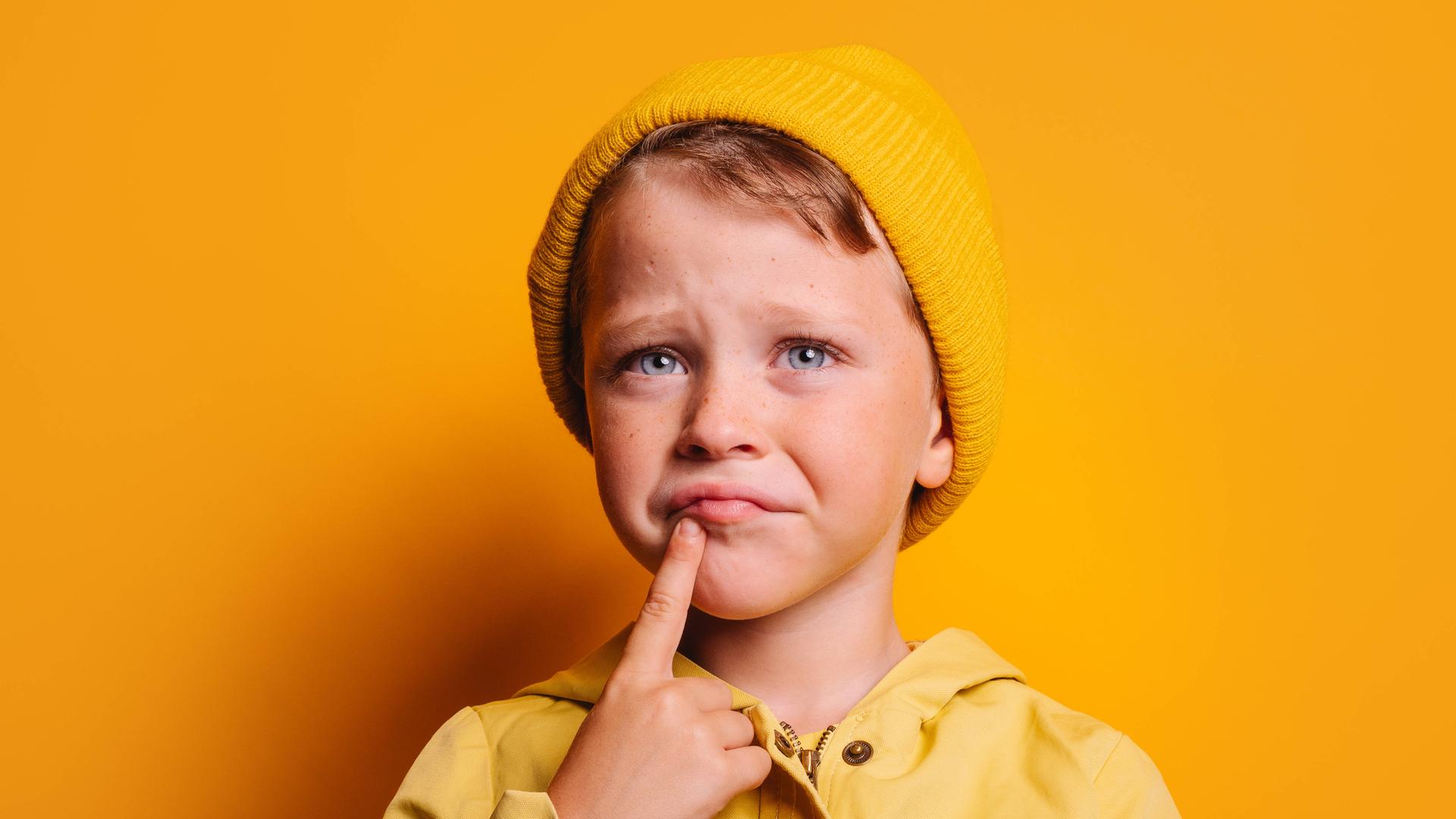 Ein Kind mit gelber Mütze vor gelbem Hintergrund mit grübelndem Gesichtsausdruck und dem Zeigefinger am Kinn