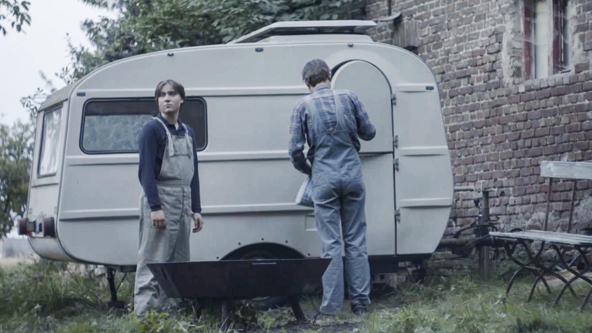 Filmstill aus dem Film "Tod meiner Jugend" zeigt eine Jungen und einen Mann mit dem Rücken vor einem alten Wohnwagen.