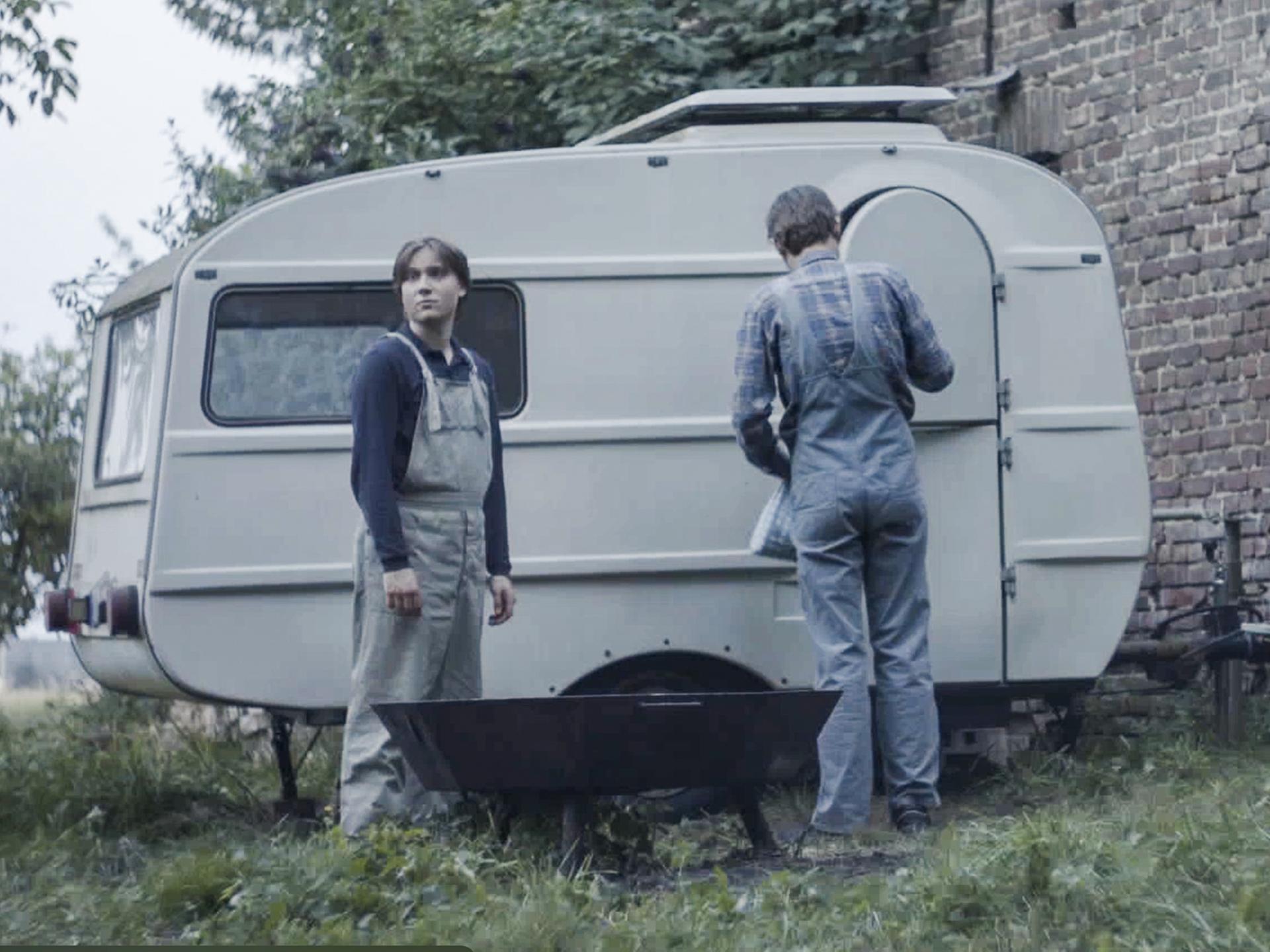 Filmstill aus dem Film "Tod meiner Jugend" zeigt eine Jungen und einen Mann mit dem Rücken vor einem alten Wohnwagen.