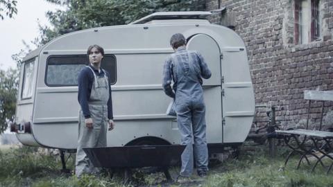 Filmstill aus dem Film "Tod meiner Jugend" zeigt eine Jungen und einen Mann mit dem Rücken vor einem alten Wohnwagen. Filmstill aus dem Film "Tod meiner Jugend" zeigt eine Jungen und einen Mann mit dem Rücken vor einem alten Wohnwagen.