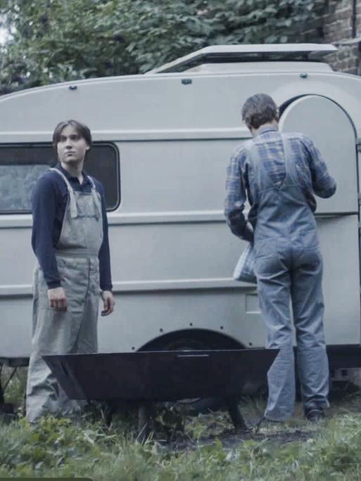 Filmstill aus dem Film "Tod meiner Jugend" zeigt eine Jungen und einen Mann mit dem Rücken vor einem alten Wohnwagen.