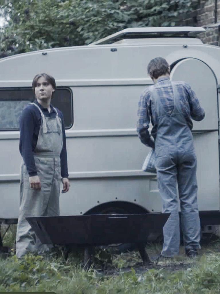 Filmstill aus dem Film "Tod meiner Jugend" zeigt eine Jungen und einen Mann mit dem Rücken vor einem alten Wohnwagen.