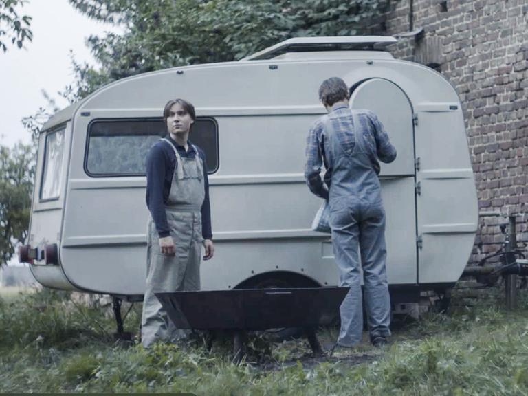 Filmstill aus dem Film "Tod meiner Jugend" zeigt eine Jungen und einen Mann mit dem Rücken vor einem alten Wohnwagen.