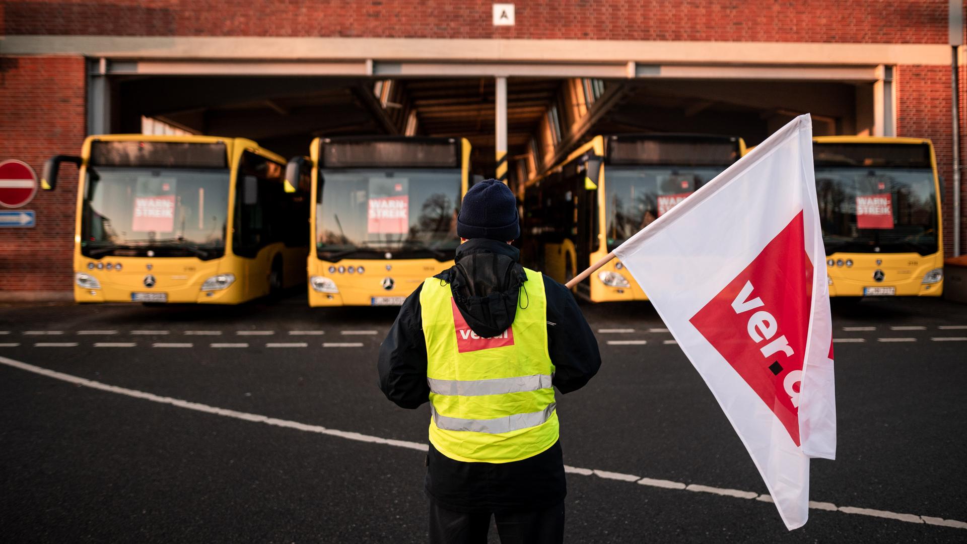 Ein Mann steht mit einer Fahne vor einigen Bussen. Der Mann hält eine Fahne der Gewerkschaft verdi in der Hand.