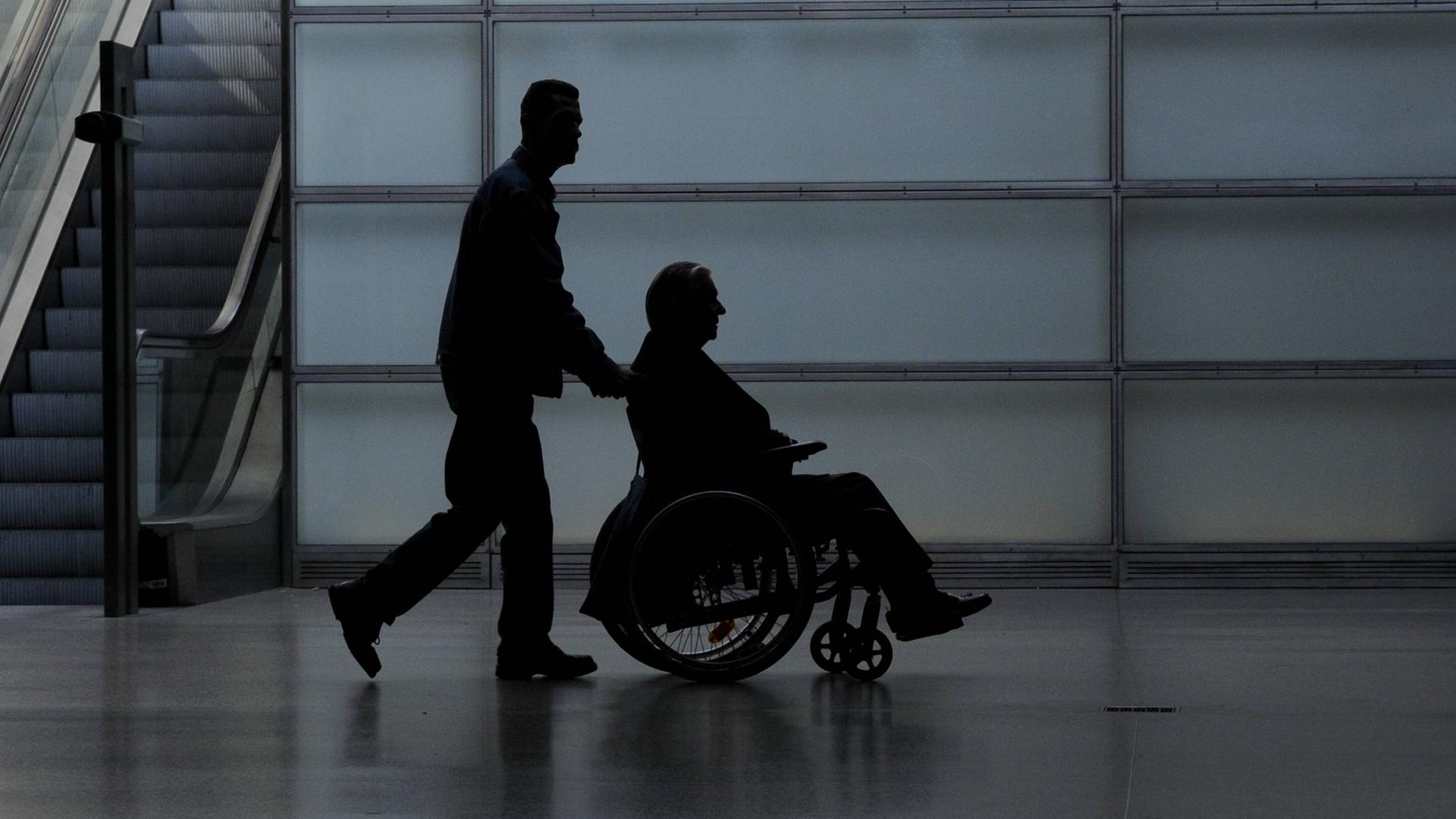 Ein junger Mann schiebt einen Rollstuhlfahrer durch eine Halle. Es könnte eine Flughafenhalle sein. Von beiden ist nur die Silhouette erkennbar. 